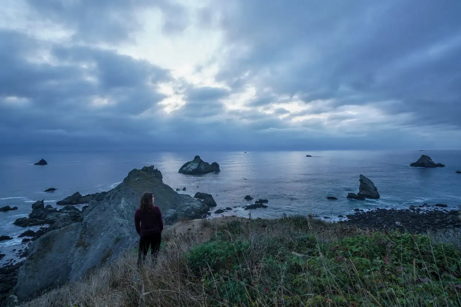 Person overlooking rocky coastline during cloudy sunset.